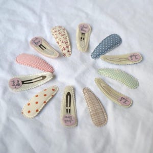 May include: A collection of fabric-covered hair clips in various patterns and colors, including polka dots, floral, and gingham. Each clip has a metal clasp and a small, round label with text. The clips are arranged in a circular pattern on a white surface.