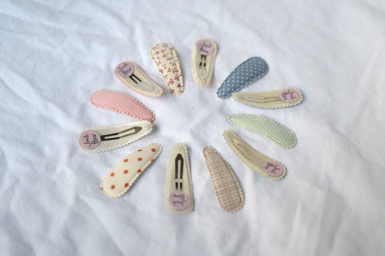 May include: A collection of fabric-covered hair clips in various patterns and colors, including polka dots, floral, and gingham. Each clip has a metal clasp and a small, round label with text. The clips are arranged in a circular pattern on a white surface.