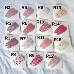 May include: A collection of fifteen fabric hair clips, each attached to a white card. The clips feature various patterns, including floral, polka dots, and gingham, in shades of pink, red, and cream. Each card is labeled with a number.