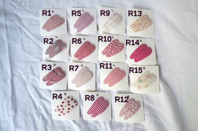 May include: A collection of fifteen fabric hair clips, each attached to a white card. The clips feature various patterns, including floral, polka dots, and gingham, in shades of pink, red, and cream. Each card is labeled with a number.