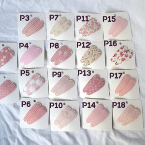 May include: A collection of eighteen hair clips, each attached to a small white card. The clips feature various patterns, including polka dots, gingham, and floral designs, in shades of pink and cream. Each card is labeled with a "P" followed by a number.