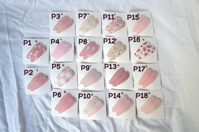 May include: A collection of eighteen hair clips, each attached to a small white card. The clips feature various patterns, including polka dots, gingham, and floral designs, in shades of pink and cream. Each card is labeled with a "P" followed by a number.