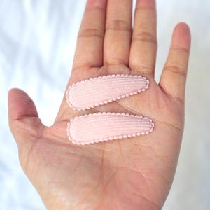 May include: Two light pink hair clips with scalloped edges are displayed on an open hand. The clips are oval-shaped and made of a textured fabric. The background is a soft, white surface.