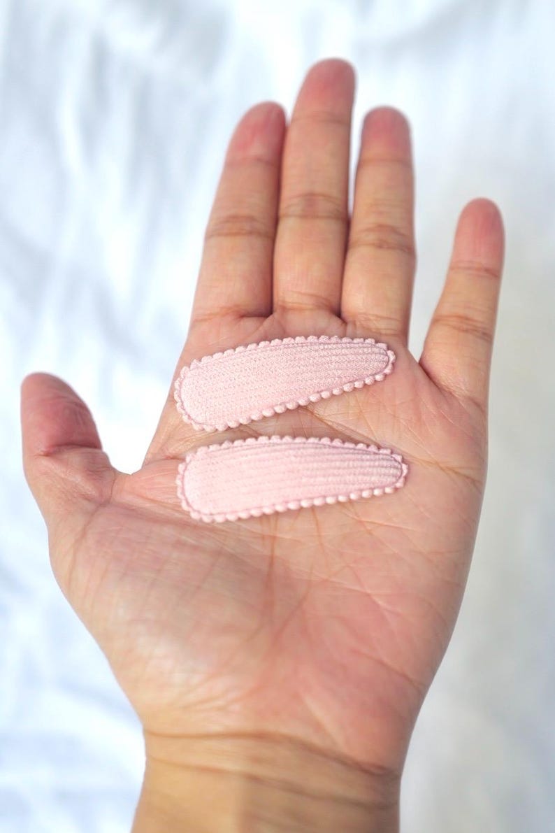 May include: Two light pink hair clips with scalloped edges are displayed on an open hand. The clips are oval-shaped and made of a textured fabric. The background is a soft, white surface.