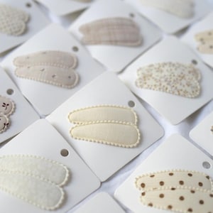 May include: An assortment of hair clips on white cards. The clips feature various patterns, including polka dots, floral prints, and textured designs. Colors include cream, beige, and brown. The clips are arranged on a white surface.
