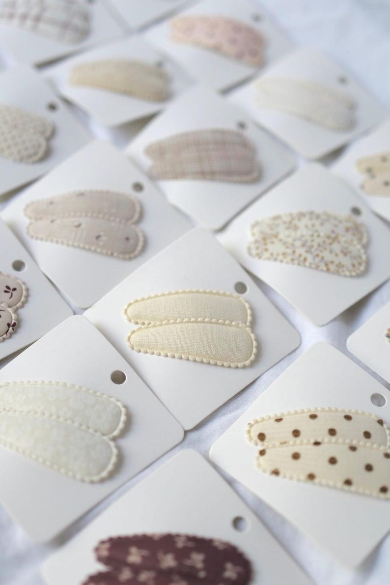 May include: An assortment of hair clips on white cards. The clips feature various patterns, including polka dots, floral prints, and textured designs. Colors include cream, beige, and brown. The clips are arranged on a white surface.