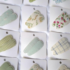 May include: Assortment of fabric-covered hair clips in various patterns, including gingham, floral, and solid colors. Each clip is attached to a white card with a small hole at the top. The clips are in shades of green, blue, and cream.