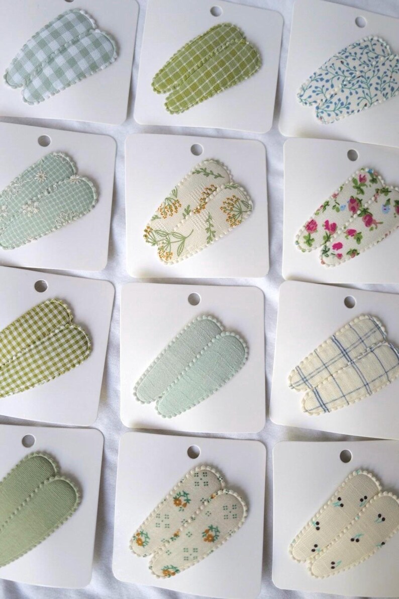 May include: Assortment of fabric-covered hair clips in various patterns, including gingham, floral, and solid colors. Each clip is attached to a white card with a small hole at the top. The clips are in shades of green, blue, and cream.