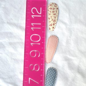 May include: Three fabric hair clips in a row, next to a pink ruler. The clips are patterned with floral, textured pink, and polka dot designs. The ruler measures up to 12 inches.
