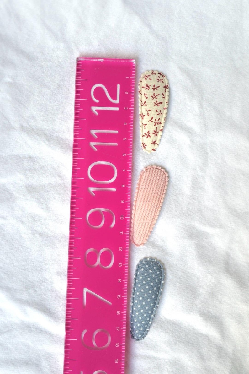 May include: Three fabric hair clips in a row, next to a pink ruler. The clips are patterned with floral, textured pink, and polka dot designs. The ruler measures up to 12 inches.