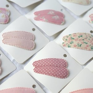 May include: Assortment of fabric-covered hair clips in various patterns, including polka dots, floral, and gingham. Each clip is mounted on a white card. The clips are primarily pink and cream colored.