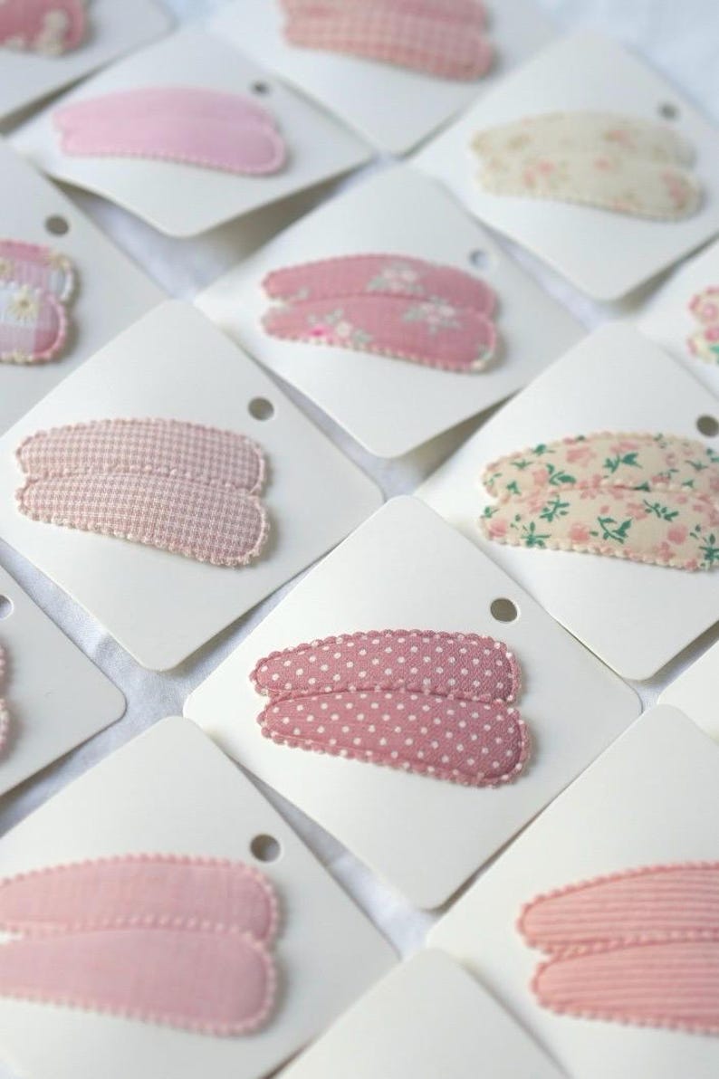 May include: Assortment of fabric-covered hair clips in various patterns, including polka dots, floral, and gingham. Each clip is mounted on a white card. The clips are primarily pink and cream colored.