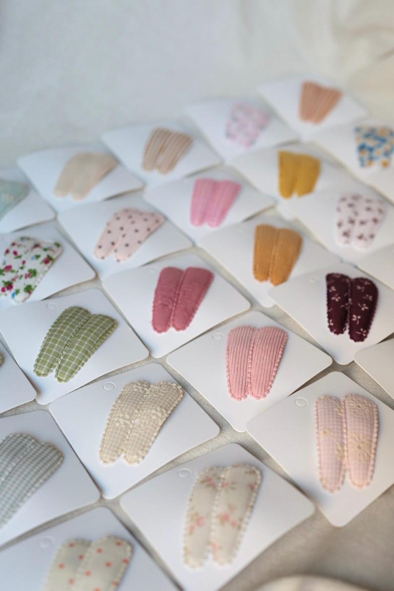 May include: A collection of hair clips in various colors and patterns, including pink, yellow, and floral designs. Each clip is displayed on a white card, creating a visually appealing arrangement. The clips are made of fabric and are likely for styling hair.