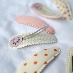 May include: A collection of hair clips in various designs and colors. The clips include a pink clip, a white clip with the text "Jreh Bow", and clips with polka dots and floral patterns. The clips are on a white surface.