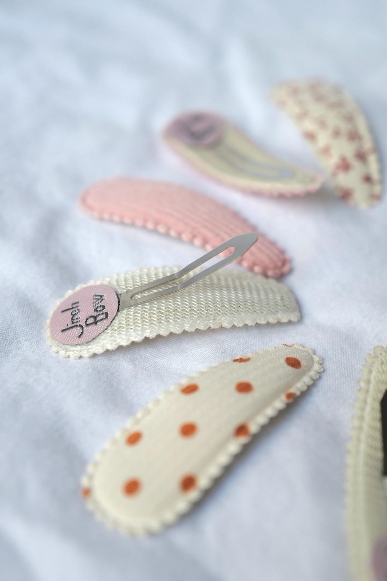 May include: A collection of hair clips in various designs and colors. The clips include a pink clip, a white clip with the text "Jreh Bow", and clips with polka dots and floral patterns. The clips are on a white surface.