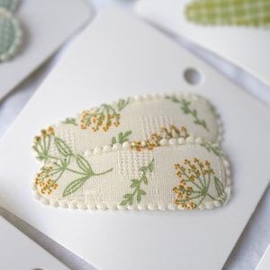 May include: Two oval-shaped hair clips with a floral pattern of green leaves and orange flowers on a cream background. The clips are on white cardstock. The edges of the clips are trimmed with a cream-colored pom-pom trim.
