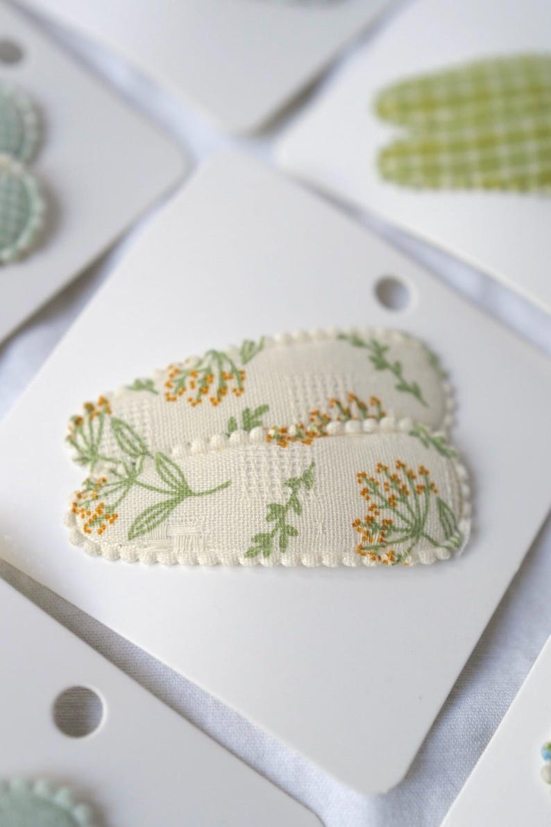 May include: Two oval-shaped hair clips with a floral pattern of green leaves and orange flowers on a cream background. The clips are on white cardstock. The edges of the clips are trimmed with a cream-colored pom-pom trim.