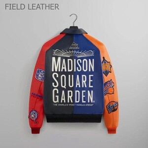 Puede incluir: Chaqueta de cuero con bloques de color con paneles rojos, azules y naranjas. La parte trasera presenta el texto "MADISON SQUARE GARDEN" en blanco. Parches con logotipos de equipos deportivos en las mangas. La chaqueta est&aacute; colgada de una percha negra.