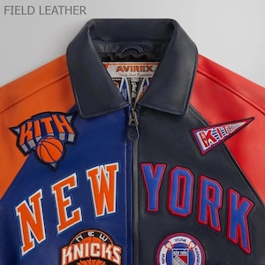 Puede incluir: Chaqueta de cuero con bloques de color en paneles naranja, azul, negro y rojo. La chaqueta presenta el texto "NEW YORK" y "KNICKS" con logotipos de baloncesto y del equipo. El cuello es azul marino y la chaqueta tiene cierre de cremallera.
