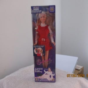 May include: A doll in a clear plastic box. The doll has blonde hair and is wearing a red dress with a white collar and belt. The box has the words "SURPRISE STYLE BABEINA" printed on it.