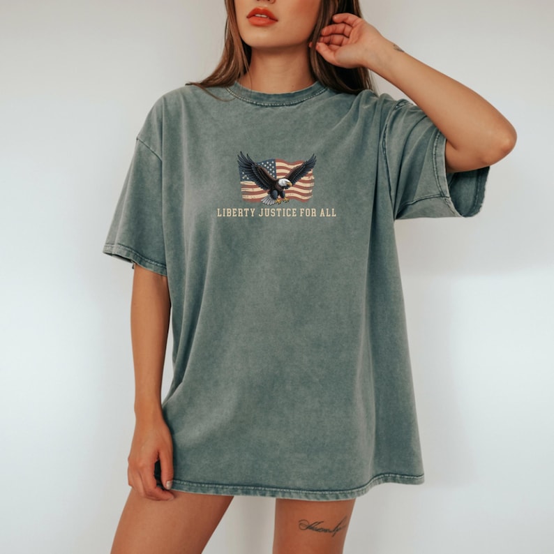 May include: A faded green t-shirt featuring an American flag design with a soaring eagle and the text "LIBERTY JUSTICE FOR ALL." The shirt has a vintage, worn look with a crew neck and short sleeves. The graphic is centered on the chest.