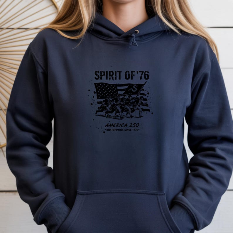 May include: Navy blue hoodie with a front graphic. The graphic features the text "SPIRIT OF '76" above an illustration of the American flag and soldiers. Below the illustration is the text "AMERICA 250" and "UNSTOPPABLE SINCE 1776".