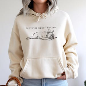May include: Cream-colored hoodie featuring a black and white illustration of a French Bulldog lying down. Above the dog is the text "CERTIFIED COUCH POTATO". The hoodie has a front pocket and a drawstring hood.