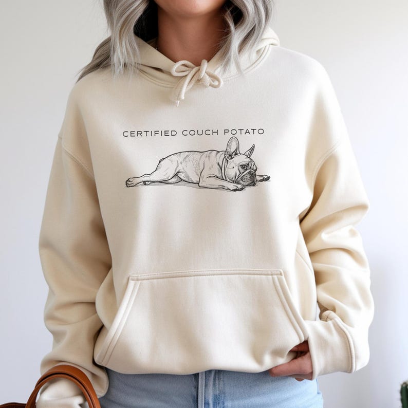 May include: Cream-colored hoodie featuring a black and white illustration of a French Bulldog lying down. Above the dog is the text "CERTIFIED COUCH POTATO". The hoodie has a front pocket and a drawstring hood.