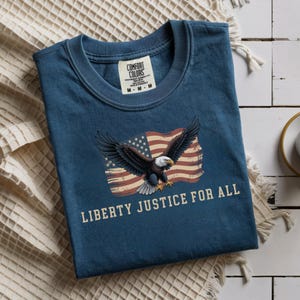 May include: A navy blue t-shirt with a graphic of an American flag and a bald eagle with outstretched wings. The text "LIBERTY JUSTICE FOR ALL" is printed below the graphic. The shirt has a crew neck and short sleeves.