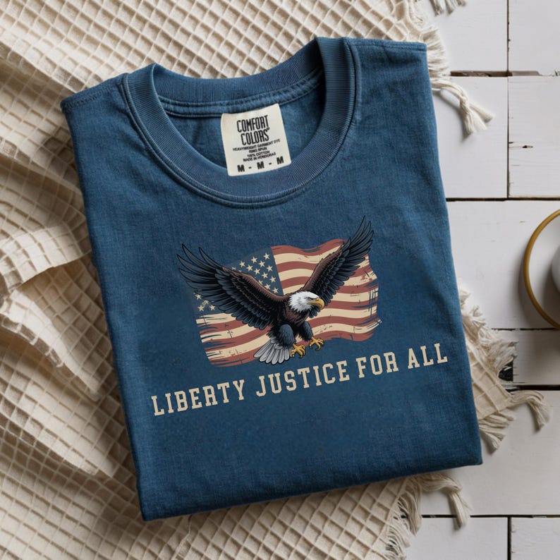 May include: A navy blue t-shirt with a graphic of an American flag and a bald eagle with outstretched wings. The text "LIBERTY JUSTICE FOR ALL" is printed below the graphic. The shirt has a crew neck and short sleeves.