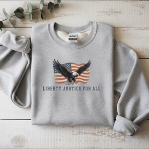 May include: A gray sweatshirt featuring an American flag and an eagle with outstretched wings. The text "LIBERTY JUSTICE FOR ALL" is printed below the graphic. The sweatshirt has a crew neck and long sleeves.