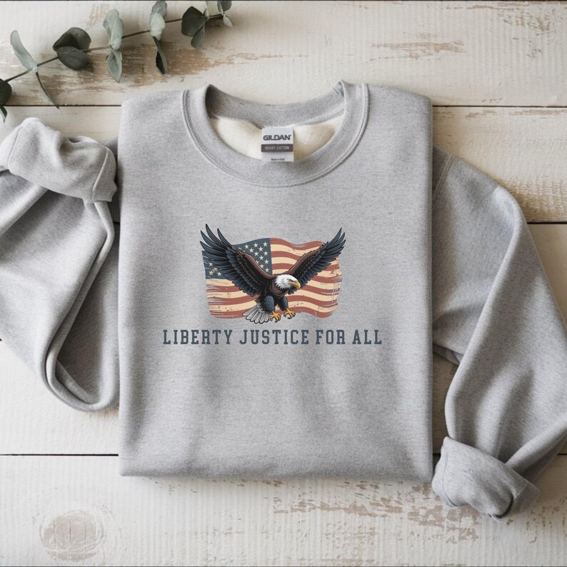 May include: A gray sweatshirt featuring an American flag and an eagle with outstretched wings. The text "LIBERTY JUSTICE FOR ALL" is printed below the graphic. The sweatshirt has a crew neck and long sleeves.
