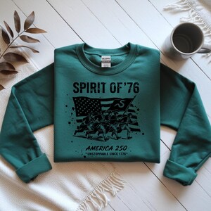 May include: A teal sweatshirt with the text "SPIRIT OF '76" above a black graphic of soldiers carrying a flag. Below the graphic, it reads "AMERICA 250 *UNSTOPPABLE SINCE 1776*". The sweatshirt has a crew neck and long sleeves.