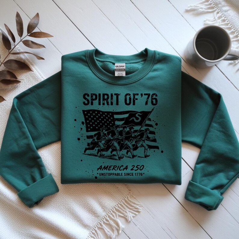 May include: A teal sweatshirt with the text "SPIRIT OF '76" above a black graphic of soldiers carrying a flag. Below the graphic, it reads "AMERICA 250 *UNSTOPPABLE SINCE 1776*". The sweatshirt has a crew neck and long sleeves.