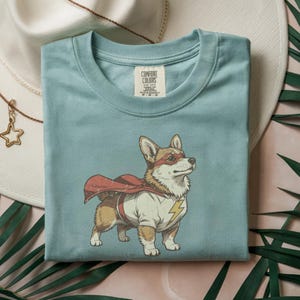 May include: A light blue t-shirt featuring a graphic of a Corgi dog dressed as a superhero with a red cape, a red eye mask, and a white shirt with a yellow lightning bolt emblem. The shirt has a Comfort Colors brand label at the inner neckline.