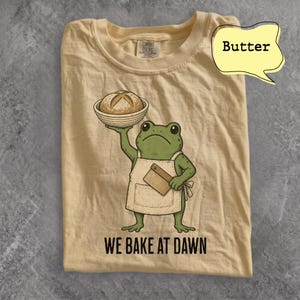 May include: A butter-colored t-shirt featuring a graphic of a green frog wearing a tan apron, holding a bowl with a loaf of bread and a small wooden tool. The text "WE BAKE AT DAWN" is printed in black below the frog. The shirt has a crew neckline.