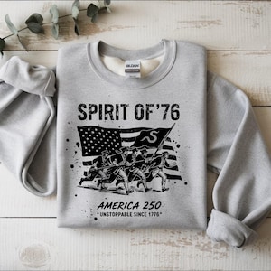 May include: A light gray sweatshirt with the text "SPIRIT OF '76" above a black and white graphic of soldiers marching in front of an American flag. Below the graphic is the text "AMERICA 250 *UNSTOPPABLE SINCE 1776*."