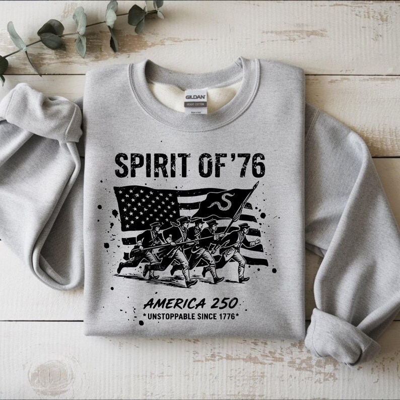 May include: A light gray sweatshirt with the text "SPIRIT OF '76" above a black and white graphic of soldiers marching in front of an American flag. Below the graphic is the text "AMERICA 250 *UNSTOPPABLE SINCE 1776*."