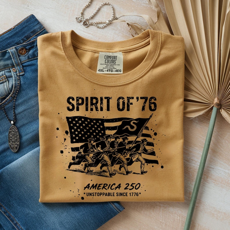 May include: Mustard-colored t-shirt with the text "SPIRIT OF '76" and an illustration of soldiers marching in front of an American flag. The text "AMERICA 250 *UNSTOPPABLE SINCE 1776*" is also printed on the shirt. The shirt is folded on top of blue jeans.
