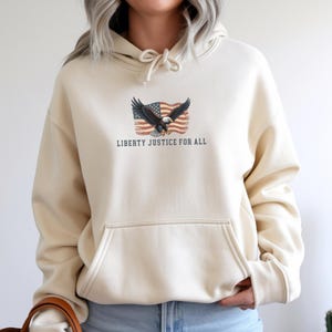 May include: Cream-colored hoodie with a front pocket and drawstring hood. The front features a graphic of an eagle with outstretched wings in front of a waving American flag. The text "LIBERTY JUSTICE FOR ALL" is printed below the graphic.