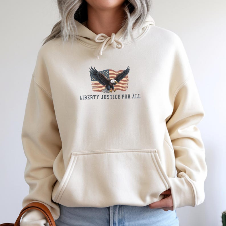 May include: Cream-colored hoodie with a front pocket and drawstring hood. The front features a graphic of an eagle with outstretched wings in front of a waving American flag. The text "LIBERTY JUSTICE FOR ALL" is printed below the graphic.