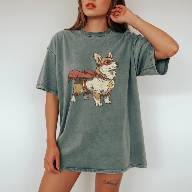 May include: A mineral-washed, slate-gray short-sleeve t-shirt featuring a graphic illustration of a Corgi dog dressed as a superhero. The dog wears a red cape, a red eye mask, and a white shirt with a yellow lightning bolt emblem on the chest.