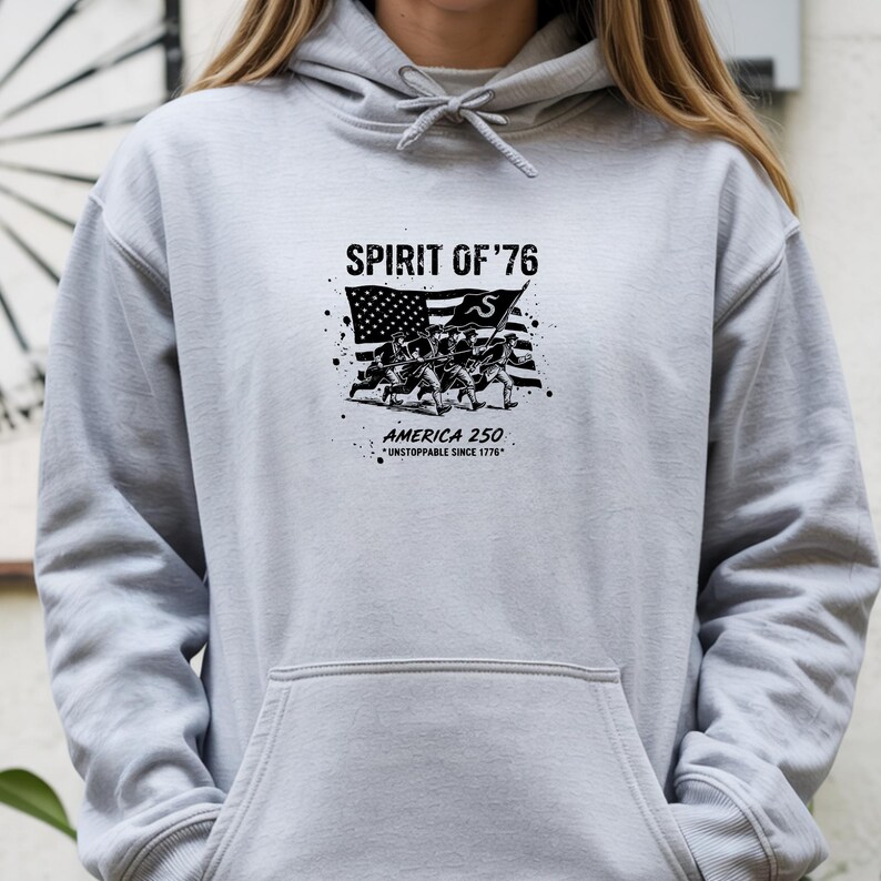 May include: A light gray hoodie featuring a black graphic with the text "SPIRIT OF '76" above an illustration of soldiers and an American flag. The text "AMERICA 250" and "UNSTOPPABLE SINCE 1776" are also displayed.