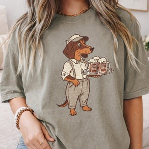 May include: Olive green t-shirt featuring a cartoon dachshund waiter wearing a cap, shirt, and suspenders. The dog is holding a tray with two mugs of root beer. The mugs have the words "Root Beer" printed on them.