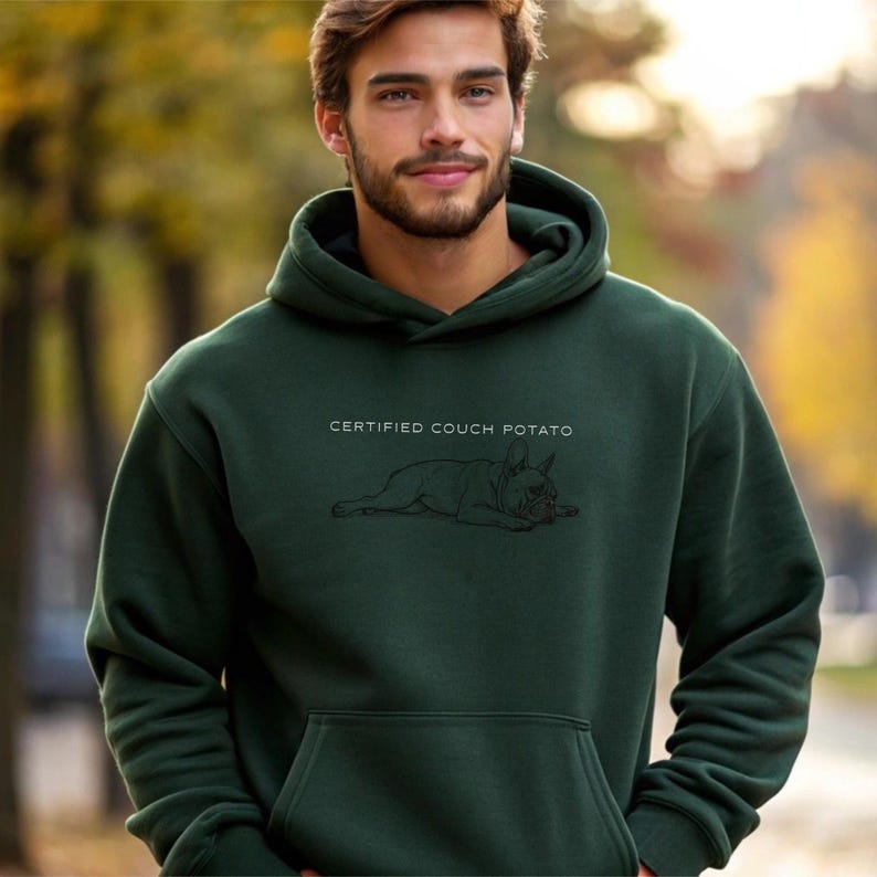 May include: Dark green hoodie with the text "CERTIFIED COUCH POTATO" above a black line drawing of a French Bulldog. The hoodie has a drawstring hood and a front pocket. The image is set outdoors.