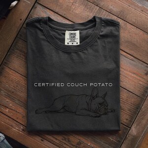 May include: Dark gray t-shirt with the words "CERTIFIED COUCH POTATO" and a line drawing of a French Bulldog. The shirt is folded on a wooden surface. The Comfort Colors tag is visible.
