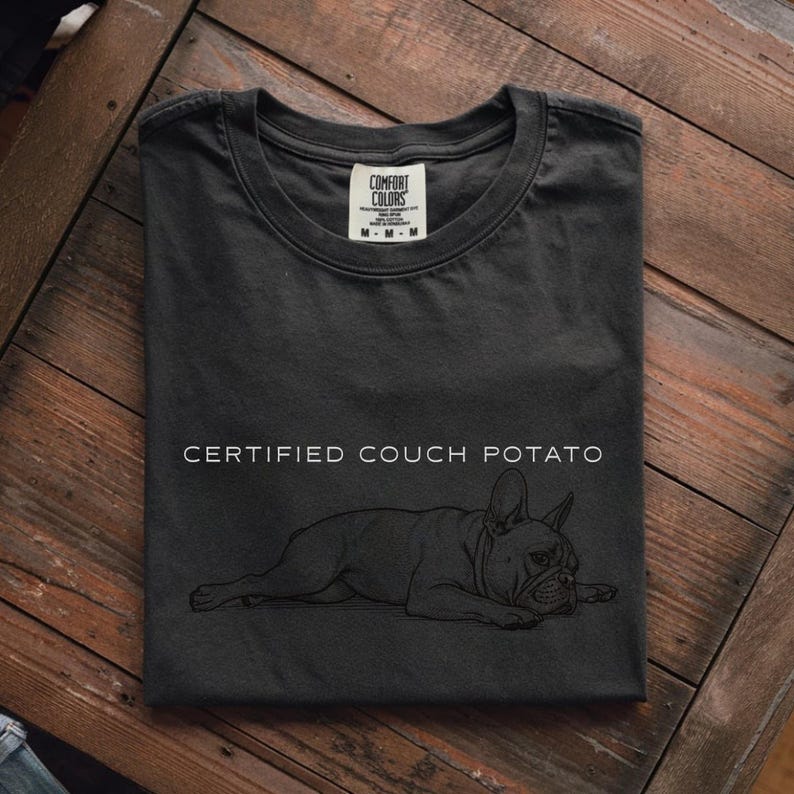 May include: Dark gray t-shirt with the words "CERTIFIED COUCH POTATO" and a line drawing of a French Bulldog. The shirt is folded on a wooden surface. The Comfort Colors tag is visible.