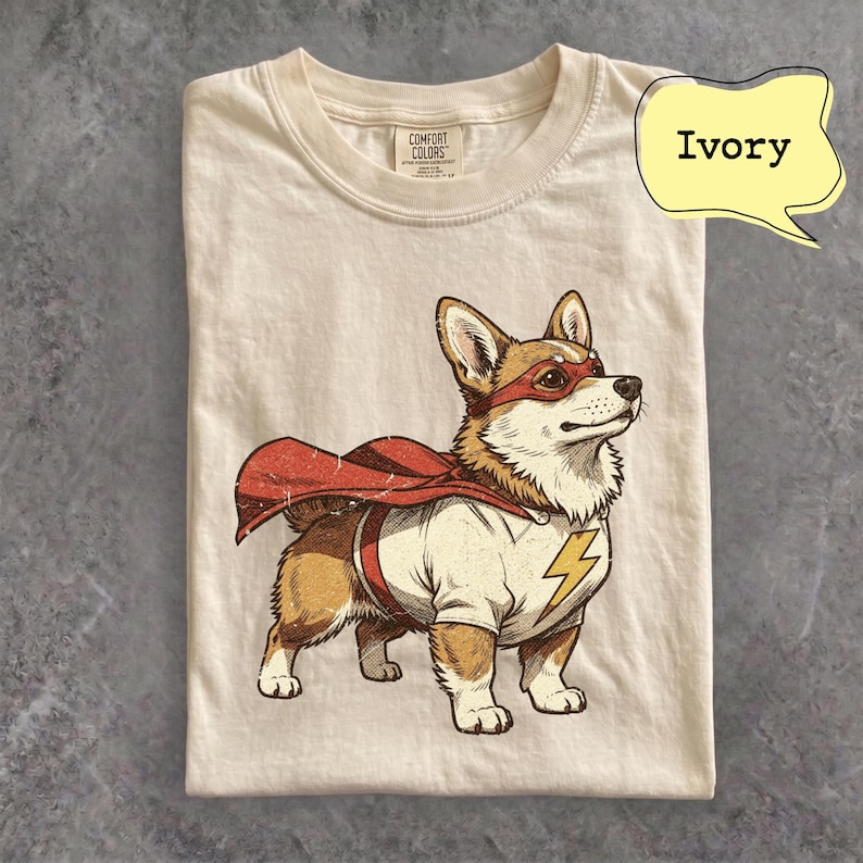 May include: An ivory short-sleeve t-shirt featuring a graphic of a Corgi dog wearing a red superhero mask and a red cape. The dog is dressed in a white shirt with a yellow lightning bolt emblem on the chest. The Comfort Colors brand tag is visible.