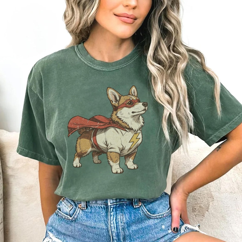 May include: A forest green short-sleeve t-shirt featuring a graphic illustration of a Corgi dog dressed as a superhero. The dog wears a red cape, a red eye mask, and a white shirt with a yellow lightning bolt emblem on the chest area.