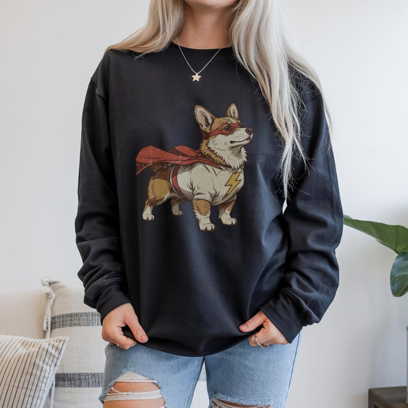 May include: A black long-sleeve sweatshirt featuring a graphic print of a brown and white Corgi dog dressed as a superhero. The dog wears a red eye mask, a red cape, and a white shirt with a yellow lightning bolt emblem on the chest area.
